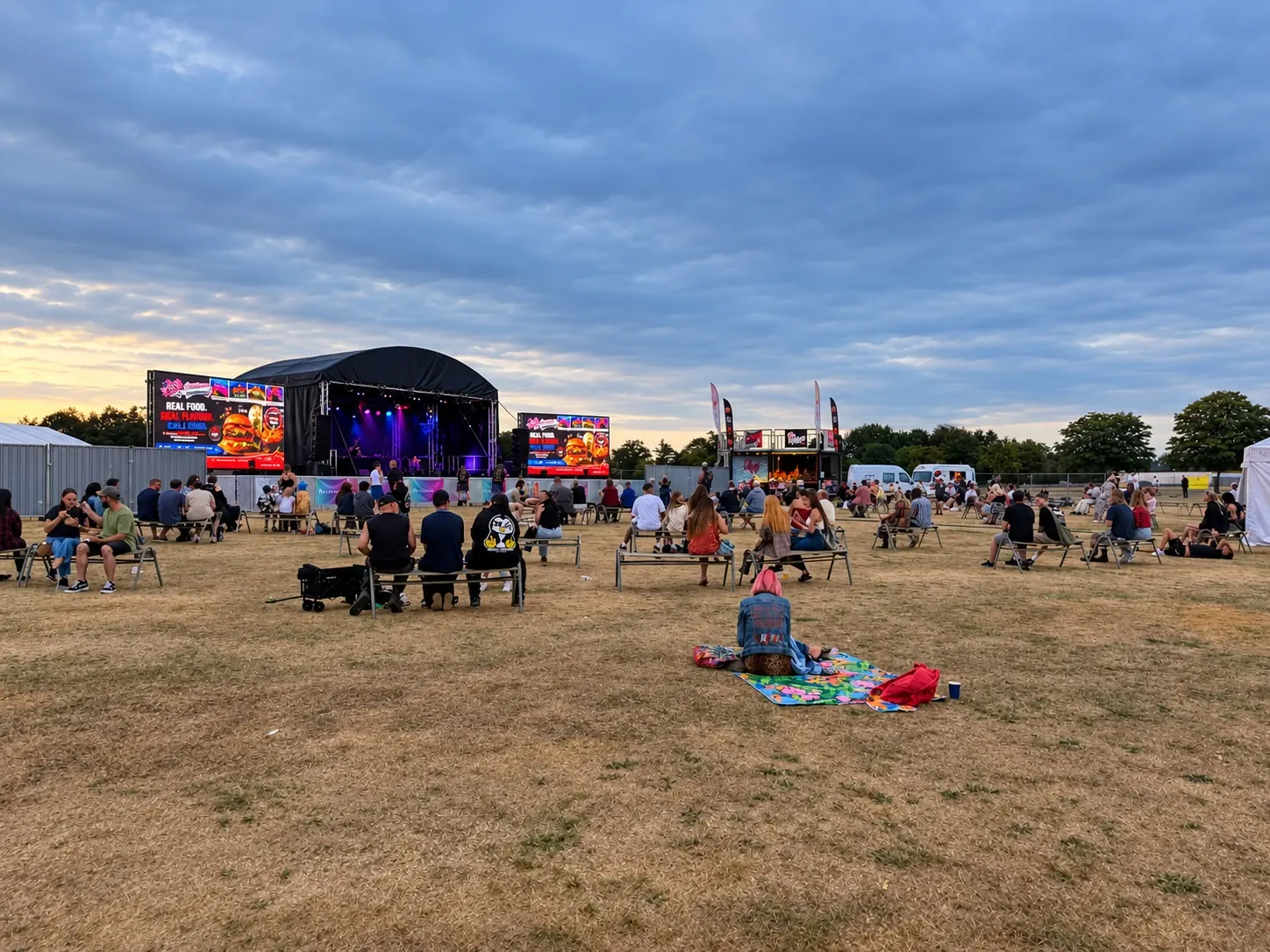 An evening crowd on benches and picnic blankets in front of a stage, with LED screens either side showing adverts